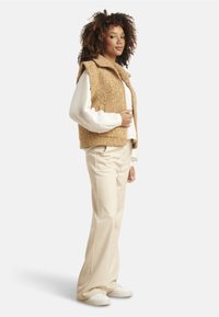 Tan textured vest with a high collar, worn over a white long-sleeve top. Off-white flared trousers and white sneakers complete the look.