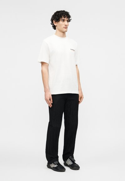 Νεαρός άνδρας με σγουρά μαλλιά που φοράει λευκό κοντομάνικο μπλουζάκι, μαύρο παντελόνι με υφή και μαύρα αθλητικά παπούτσια με λευκές ρίγες, όρθιος.