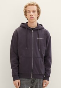 Dunkelblauer Reißverschluss-Hoodie aus Baumwollmischung, mit einer Fronttasche, einer verstellbaren Kapuze und Logo auf der linken Brust. Glatte Textur.