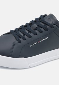 Navy modré kožené tenisky s bílými gumovými podrážkami, se šňůrkami, texturovaným povrchem a logem na boku. Jednoduchý a moderní design.