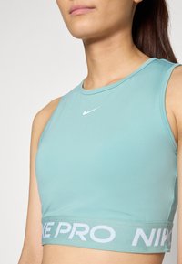 Svijetloplava Nike Pro sportski grudnjak s okruglim izrezom, širokim bijelim logotipom na dnu i glatkom, rastezljivom tkaninom.