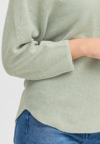 Maglione lavorato a maglia verde chiaro con un motivo testurizzato, dotato di maniche a tre quarti e orlo curvo, abbinato a jeans blu.