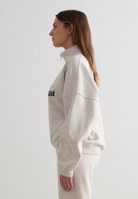 Sudadera beige claro con cremallera, mangas largas, puños acanalados y bolsillos laterales. Texto negro audaz en el frente. Tela suave y texturizada.