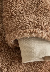 Braunes Plüschgewebe mit einer strukturierten, fischigen Oberfläche. An der Kante ist eine hellere beige, glatte Baumwollschicht sichtbar, die einen Kontrast zwischen den Materialien zeigt.