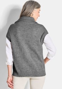 Pull en maille sans manches gris avec un col montant, bords côtelés et une finition texturée, porté par-dessus une chemise blanche à manches longues.