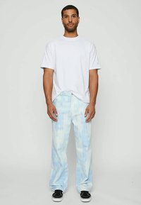 Homem de pé, vestindo uma t-shirt branca simples, jeans padrão azul claro e ténis pretos, diante de um fundo claro e uniforme.