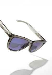 Ochelari de soare supradimensionați cu tentă gri și lentile mov, având un cadru transparent și logo-ul "HAWKERS" pe lateral.