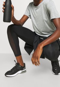 Leggings deportivas negras con detalles de malla, camiseta gris de Nike, zapatillas negras y una botella de agua negra, posicionados en una postura de cuclillas.