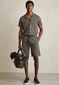 Rövid ujjú, galléros ing és nadrág szett geometrikus fekete-fehér mintával. A modell egy sötét sporttáskát tart, és sötét sneakers-t visel.