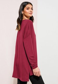 Burgundy long-sleeve top with a relaxed fit, side slits, and soft fabric, paired with black fitted pants.