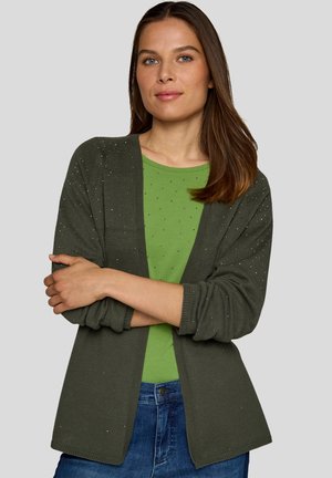 Woman with long brown hair wearing an olive green knit cardigan over a green top with small dots and blue jeans, arms crossed.
