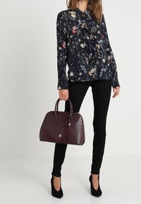 Blusa floreale blu navy con davanti arricciato, pantaloni skinny neri e borsa a mano in crosta di coccodrillo borgogna scuro con hardware e manici dorati.