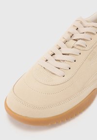Beige semsket sneaker med rund tå, flate snører og gummisåle. Har stikninger og et preget logo på siden.