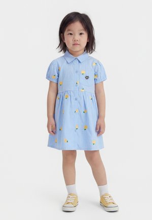 Μικρό κορίτσι που φοράει ανοιχτό μπλε φόρεμα με κίτρινα μοτίβα φρούτων, λευκές κάλτσες και κίτρινα αθλητικά παπούτσια, στέκεται μπροστά σε απλό λευκό φόντο.