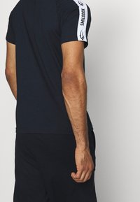 Svart idrottströja med korta ärmar, med vita accentstrimmor och logotyp på axeln. Mjuk tygkvalitet med en figurnära design.