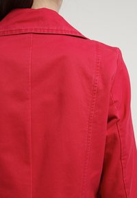 Chaqueta roja de algodón con cuello de solapa, con textura suave y costuras visibles a lo largo de las uniones. Vista trasera que muestra un diseño entallado.