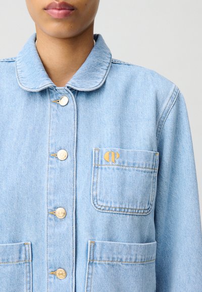 Veste en denim bleu clair avec un col arrondi, des boutons dorés, deux poches avant et un logo brodé sur la poche poitrine.