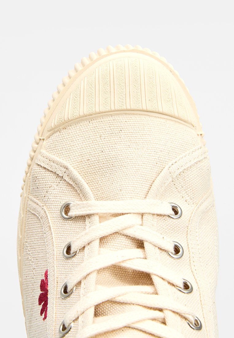 Baskets en toile beige avec un embout et une semelle en caoutchouc, œillets en métal, lacets blancs et une petite fleur rouge brodée sur le côté.