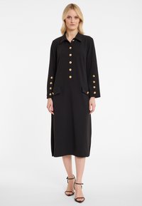 Schwarzes Kleid mit Kragen, goldenen Knopfverschlüssen und Taschen; lange Ärmel mit goldenen Knopfakzenten. Glatte Textur, Midi-Länge.