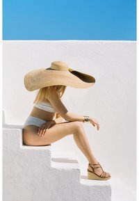 Cappello di paglia a falda larga, bikini bianco, seduta su gradini bianchi, con sandali con zeppa beige e cinturini, su uno sfondo di cielo azzurro.