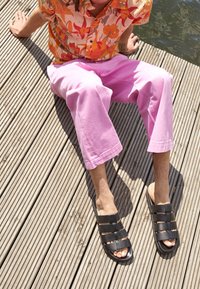 Sandali neri con cinturini e design elegante, con punta aperta, abbinati a pantaloni larghi rosa, su una superficie in legno.