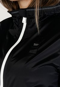 Giacca nera in nylon con colletto alto, cerniera bianca e un piccolo tag con marchio sul petto. Texture liscia con cuciture a contrasto.