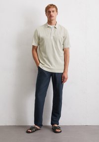 Jasnozielona koszulka polo z krótkim rękawem, z kołnierzykiem i zapięciem na trzy guziki. W zestawie z ciemnogranatowymi spodniami i czarnymi sandałami.