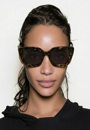 Femme aux cheveux noirs mouillés portant de grandes lunettes de soleil rectangulaires à motif écaille de tortue et un haut noir sur un fond clair uni.