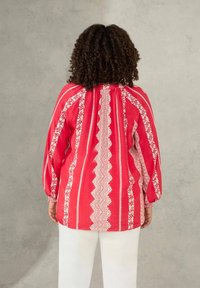 Rote Bluse mit langen Ärmeln, floralen und geometrischen weißen Mustern, gerafftem Rücken und elastischen Bündchen. Kombiniert mit weißen Hosen.