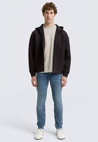 Fekete, cipzáras kapucnis pulóver laza szabással, világos bézs póló fölött viselve. Kenguruzsebekkel rendelkezik, világoskék farmerrel és fehér tornacipővel párosítva.