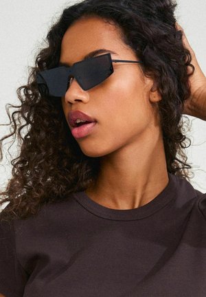 Black angular sunglasses with wire frame. Textured dark t-shirt, close-up of model's face features curly hair and neutral lip color.