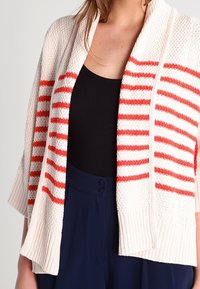 Pletený cardigan v krémové barvě s červenými horizontálními pruhy, s šálovým límcem, otevřeným předním dílem a žebrovanými manžetami a lemem. Měkká textura.
