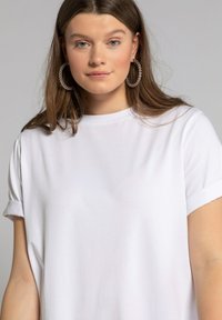 Biely bavlnený tričko s rolovanými rukávmi a klasickým okrúhlym výstrihom. Modelka doplnila svoj vzhľad veľkými striebornými kruhovými náušnicami.