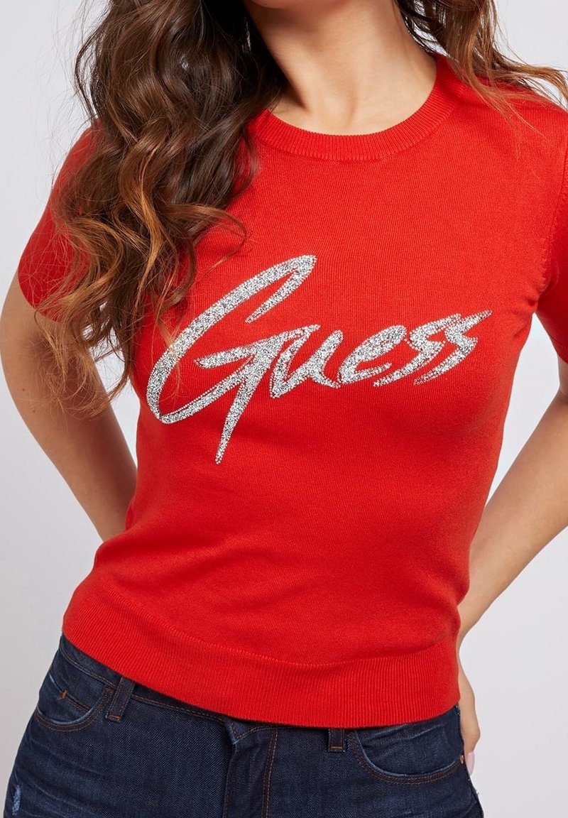 Femme portant un t-shirt rouge à manches courtes avec "Guess" en grandes lettres argentées, assorti à un jean bleu foncé, vue de la poitrine aux hanches.
