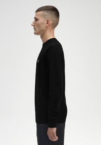 Schwarzer Pullover mit langen Ärmeln, runder Ausschnitt und gerippten Bündchen. Enthält ein kleines Emblem auf der Brust und hat ein schlichtes, körperbetontes Design.