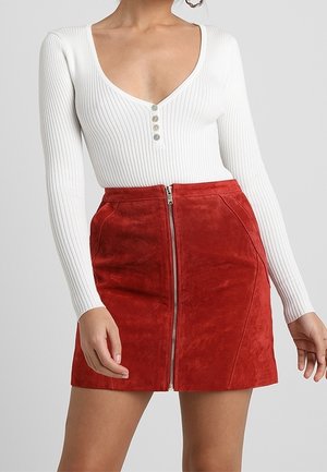 Femme portant un haut blanc côtelé à manches longues avec des boutons et une mini-jupe rouge en daim avec une fermeture éclair à l'avant.