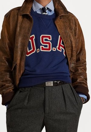 Personne portant une veste en cuir marron sur un sweat bleu USA, une chemise rayée avec une cravate tricotée, un pantalon plissé gris et une ceinture noire.