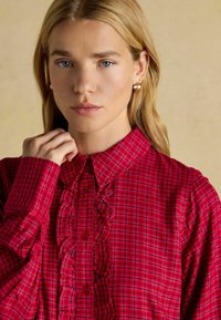 Red plaid shirt with a button-up front, featuring ruffled accents along the placket and a classic collar. Soft, cotton-like texture.