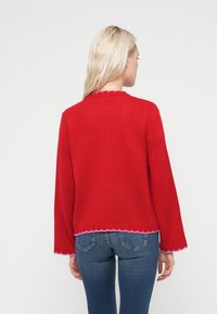 Κόκκινο πλεκτό πουλόβερ με λαιμόκοψη και τελείωμα σε σχήμα κυματιστού άκρου. Διαθέτει μακριά μανίκια και αντίθετη ροζ ραφή στα άκρα.