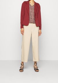 Blazer sur mesure bordeaux porté sur un chemisier à motifs, associé à un pantalon large beige et des sandales noires à lanières. Textures lisses mises en avant.