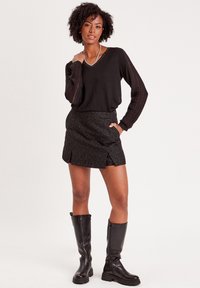 Schwarzer Strickpullover mit strukturierten Ärmeln, kombiniert mit einem glitzernden schwarzen Minirock mit einem Schlitz vorne und kniehohen schwarzen Stiefeln.