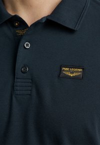 Dunkelblaues Polohemd mit einem Kragen, zwei schwarzen Knöpfen und einem PME Legend-Label mit gelben Akzenten oben links. Strukturierter Stoff.