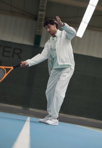 Conjunto de ténis leve em verde menta claro, composto por uma jaqueta com fecho texturizada e calças combinando. Segura uma raquete laranja numa superfície de court azul.