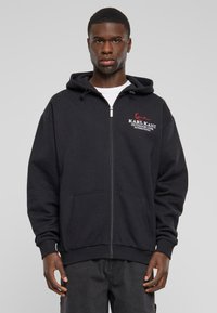 Sudadera con cremallera negra hecha de tela suave, con un bolsillo tipo canguro y capucha con cordón. Logotipo bordado en rojo y blanco en el pecho.