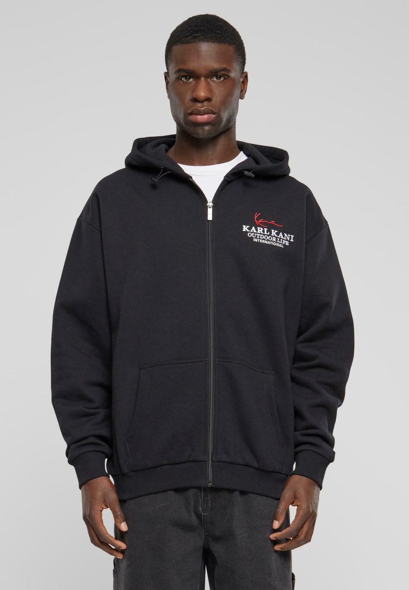 Sudadera con cremallera negra hecha de tela suave, con un bolsillo tipo canguro y capucha con cordón. Logotipo bordado en rojo y blanco en el pecho.