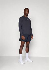 Tumesinise värviga pika varrukaga ülaosa logoga, kombineeritud tumedate pükstega. Modell kannab valgeid sportkingi ja sokke. Sile kangas, spordiriietus.