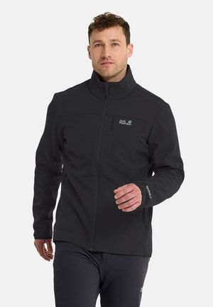 Man draagt een zwarte Jack Wolfskin jas met rits en hoge kraag, loopt naar voren tegen een effen lichte achtergrond.