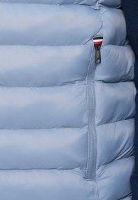 Világoskék puffert mellény vízszintes steppeléssel, cipzáras zsebbel, amely fekete, piros és fehér díszítéssel van kiemelve. Simaságú anyag.