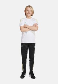 Witte sportshirt met korte mouwen, voorzien van lichtblauwe accenten en rode stiksels, gecombineerd met zwarte broek met gele strepen.