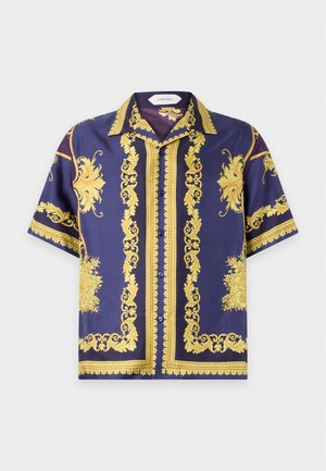 Homme portant une chemise violette à manches courtes avec des motifs baroques dorés, un pantalon noir, des chaussures noires aux bouts dorés et un collier en or, debout immobile.